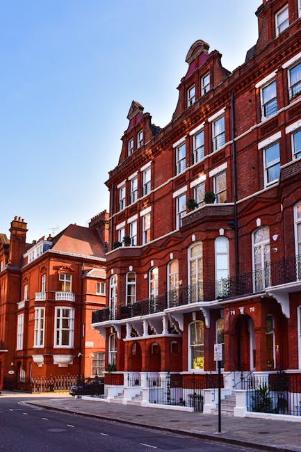 Exterior view of a row of red-brick residential buildings on Kensington High Street, featuring large sash windows, decorative white trim, and balconies with black wrought iron railings. The buildings are well-maintained with clean brickwork and tidy window areas. The street in front has a smooth asphalt surface with designated parking spaces and a clear sidewalk. Bright daylight illuminates the scene, highlighting the building's architectural details. Cleaners West Kensington specializes in surface cleaning, deep cleaning, and sanitisation for residential properties, ensuring each element of the exterior remains pristine and well-maintained, as seen in this tidy urban streetscape.