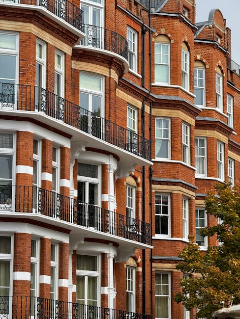 A close-up view of a multi-story residential building with red brick facade, white decorative accents, and black metal balconies. The building features large rectangular windows with white trim, some with open blinds. The balconies have black railings and are supported by white columns. The overall appearance is clean and well-maintained, with natural daylight illuminating the facade. A tree with green and yellow leaves is visible in the lower right corner, contributing to a tidy urban streetscape. This image highlights the exterior condition of a typical London apartment building, illustrating the importance of surface cleaning and maintenance, which is a core service offered by Cleaners West Kensington for end-of-tenancy and domestic cleaning needs.