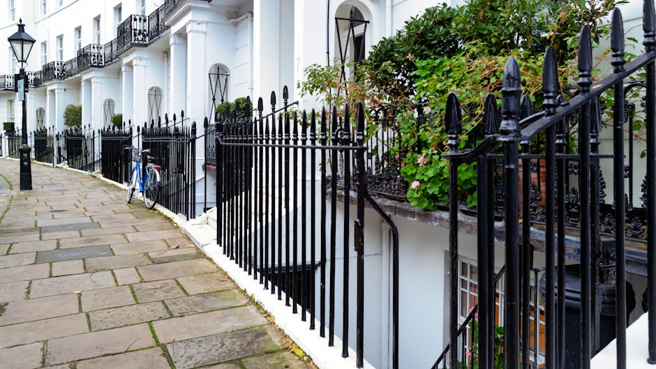Exterior view of a row of red-brick residential buildings on Kensington High Street, featuring large sash windows, decorative white trim, and balconies with black wrought iron railings. The buildings are well-maintained with clean brickwork and tidy window areas. The street in front has a smooth asphalt surface with designated parking spaces and a clear sidewalk. Bright daylight illuminates the scene, highlighting the building's architectural details. Cleaners West Kensington specializes in surface cleaning, deep cleaning, and sanitisation for residential properties, ensuring each element of the exterior remains pristine and well-maintained, as seen in this tidy urban streetscape.